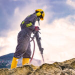 Worker with helmet and protective suit using a drilling machine on top of a large rock
